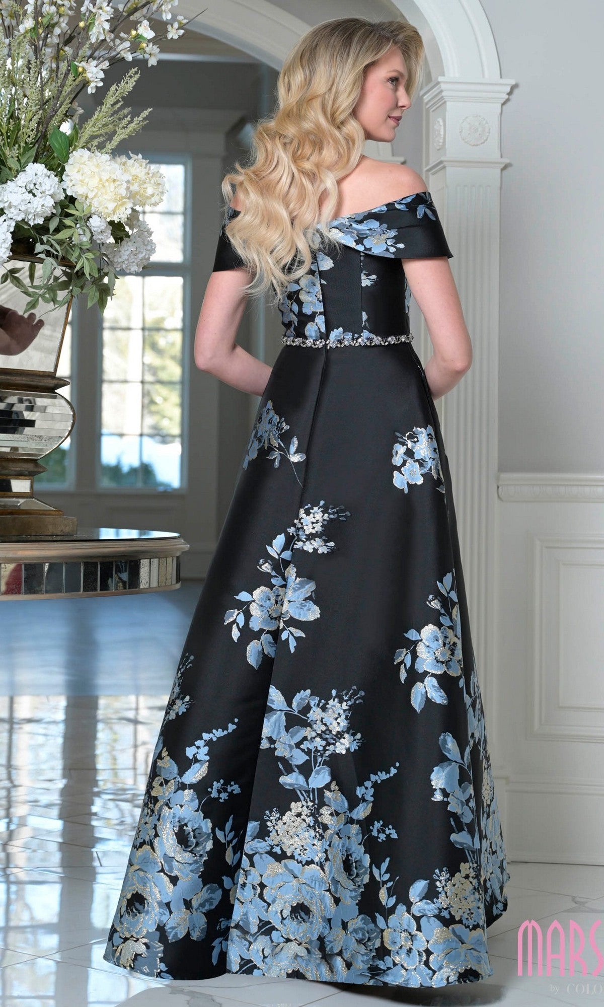 A woman with long blonde hair stands indoors, back to the camera, wearing the Marsoni MV1402—an off-the-shoulder long formal dress featuring blue floral jacquard patterns.